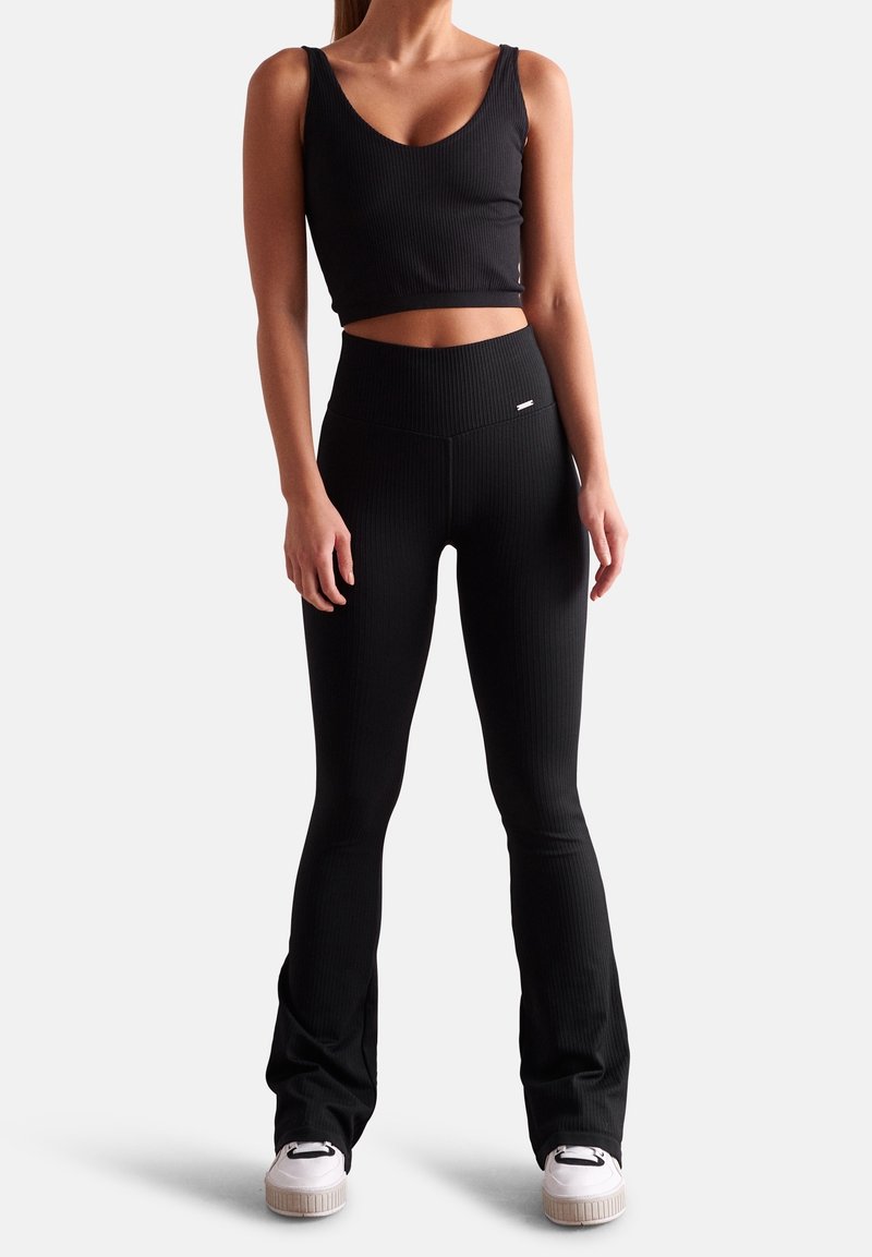 Top corto negro de canalé con tirantes anchos combinado con leggings negros acampanados; ambos hechos de una tela elástica. Detalle del logotipo en blanco en los leggings.