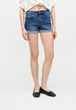 Vrouw die blauw denim shorts met omgezoomde rand en drie knopen aan de voorkant draagt, een wit cropped blouse met bloemenpatroon en zwarte balletschoentjes.