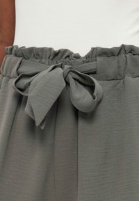 Pantalon gris taille haute avec une ceinture élastique froncée et une ceinture en tissu nouée en un nœud, porté avec un haut blanc.