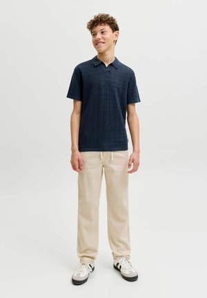 Teismeline lokkis juustega, kandes tumelilla lühikeste varrukatega polo särki, beežid püksid ja valged tossud, seisab ja naeratab.