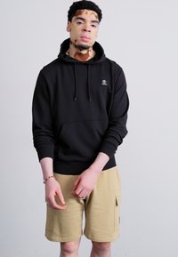 Black hoodie with a front pocket and logo, paired with tan shorts featuring side pockets. Fabric appears smooth with a relaxed fit.
