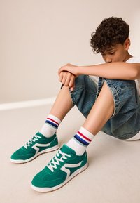 Baskets en daim vert avec des accents blancs et des lacets blancs. Portées avec des chaussettes blanches ornées de rayures rouges et bleues, associées à un short en jean.