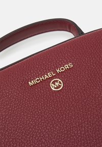 Bordeaux gekleurde leren handtas met textuur, voorzien van een bovenhandvat, gouden "MICHAEL KORS" logo en ronde gouden accent met initialen.