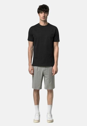 Uomo in piedi che indossa una maglietta nera a maniche corte, pantaloncini grigio chiaro al ginocchio, calzini bianchi e scarpe da ginnastica bianche su uno sfondo semplice.