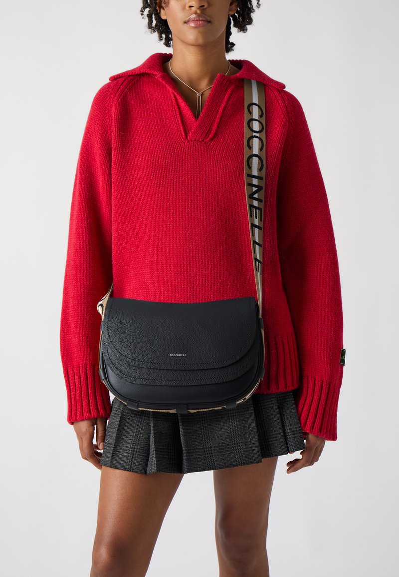 Sort læder crossover taske med en buet klap, en brun accentrem med "COCCINELLE" i sort. Båret over en rød sweater og et ternet nederdel.