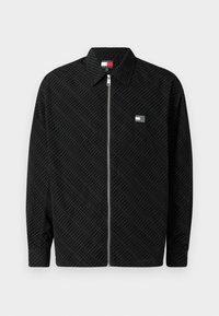 Juodas zip-up bliuzonas su tekstūruotu raštu. Yra apykaklė ir mažas logotipo lipdukas kairiajame krūtinės šone. Ilgos rankovės su tiesiu kirpimu.