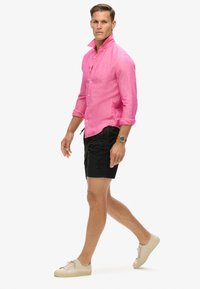 Camicia rosa a maniche lunghe, con risvolti, abbinata a pantaloni corti neri con logo e sneaker casual beige.
