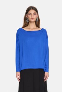 Niebieska bluzka z długim rękawem i szerokim dekoltem, wykonana z miękkiego materiału. Posiada opadające ramiona i luźny krój, zestawiona z czarną spódnicą.