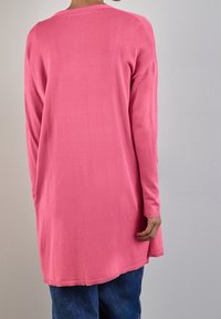 Dlouhý svetr s dlouhým rukávem, měkká pletenina, jasně růžová barva, kulatý výstřih, boční rozparky na lemu, nošený přes džíny.