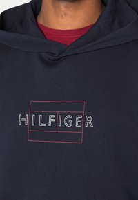 Felpa blu marina in cotone con logo grafico frontale. Presenta un cappuccio con cordino e una texture a costine. Indossata con una maglietta rossa sotto.