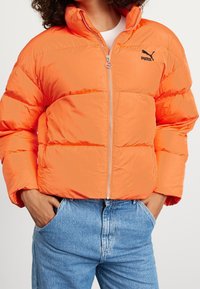 Giacca a vento arancione brillante, con chiusura a zip, colletto alto e un logo nero. Indossata sopra jeans in denim azzurro chiaro.