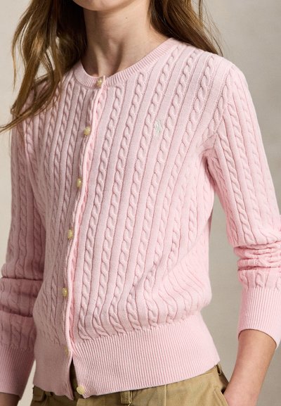 Nő világos rózsaszín kötött kardigánt visel gyöngy gombokkal és bézs nadrággal, vállától a csípőig látható, semleges háttér előtt.