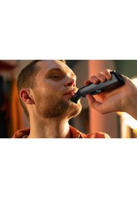 Black beard trimmer with a green indicator light, held to a man’s face. Features a sleek design and a textured grip for easy handling.
