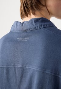 Chemise à col bleu foncé en tissu texturé avec un logo discret au dos. Présente une coupe décontractée et des détails de couture visibles.