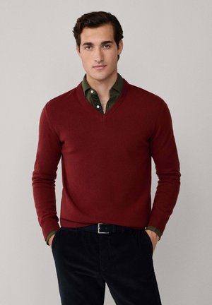 Roter V-Ausschnitt-Pullover aus weichem Material, getragen über einem dunkelgrünen Hemd mit Kragen. Das Outfit hat eng anliegende Ärmel und eine glatte Oberfläche.