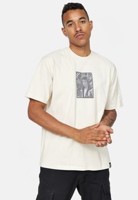 Romige katoenen t-shirt met een zwart-wit fotoafdruk in het midden, ronde ronde halslijn en korte mouwen. Bevat een klein logotag.