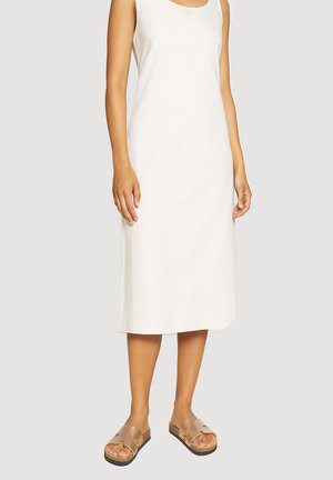 Donna che indossa un vestito midi bianco senza maniche e sandali beige slip-on, in piedi davanti a uno sfondo grigio chiaro uniforme.