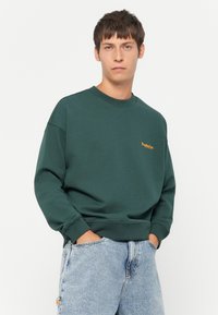 Dark green sweatshirt with a crew neck, featuring an orange brand logo on the chest. Long sleeves, relaxed fit, soft texture.