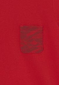 Punainen kangas, jossa on teksturoitu tasku, jossa on vuoristoteema saman sävyisessä punaisessa. Tasainen pinta, yksinkertainen muotoilu, ei näkyvää metalliosaa.