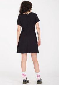Abito nero a maniche corte con corpetto aderente e gonna svasata; materiale in cotone morbido; chiusura con zip sul retro; indossato con calze bianche decorate con fiamme rosa.