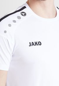 Biała koszulka sportowa z krótkim rękawem z czarnymi akcentami na ramionach, logo na piersi oraz teksturowany detal wzdłuż szwu ramienia.