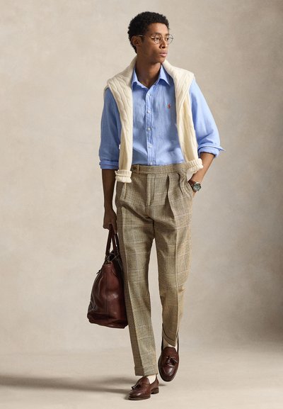 Ανοιχτό μπλε πουκάμισο με κουμπιά, εκρού πλεκτό γιλέκο, καρό παντελόνι, καφέ μοκασίνια, κρατώντας καφέ δερμάτινη τσάντα· στρογγυλό ρολόι στον καρπό.