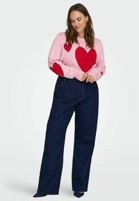 Růžový svetr s velkými červenými vzory ve tvaru srdce, spárovaný s tmavě modrými džínovými kalhotami s vysokým pasem. Hladká textura s žebrovanými manžetami a pasem.