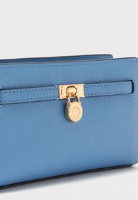 Blauwe leren portemonnee met een gestructureerde afwerking, voorzien van een gouden metalen sluiting en een ronde logo-tag. Compact, rechthoekig ontwerp, minimalistische accenten.