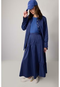 Blauwe blouse met knopen, bijpassende blauwe top en lange blauwe rok met ruche-details. Draag het met witte sneakers en een blauwe pet.