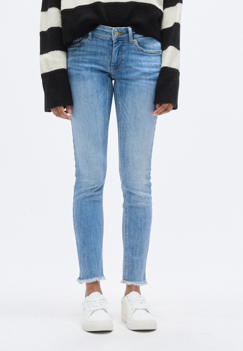 Γαλάζια στενή τζιν παντελόνα με ξεθωριασμένο φινίρισμα και ακατέργαστο τελείωμα, συνδυασμένη με λευκά αθλητικά παπούτσια. Το ύφασμα μοιάζει με τζιν με ελάχιστη υφή.