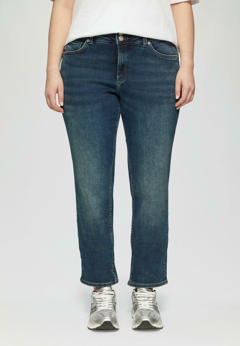 Jean en denim bleu foncé avec une coupe droite, comportant des poches avant et un zip à boutons, associés à des baskets de sport argentées.