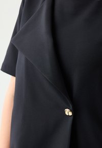 Blusa nera in tessuto liscio con colletto drappeggiato, caratterizzata da una chiusura frontale con bottone dorato. Maniche corte e design elegante.