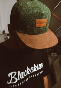 Zöld textúrájú sapka barna velúr karimával, rajta egy téglalap alakú bőrkiemelés "Blackskies" felirattal. Fekete póló fehér logóval látható alatta.