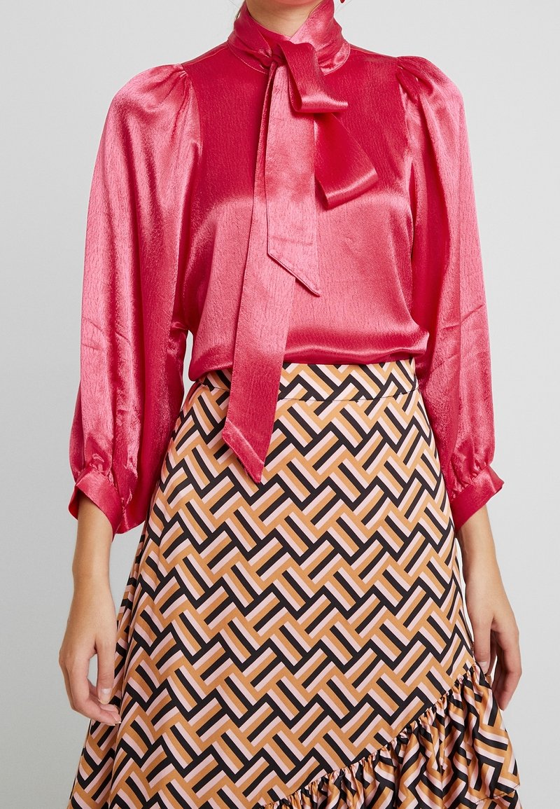 Blusa di raso rosa con maniche a sbuffo lunghe e un fiocco al colletto, abbinata a una gonne midi stampata con forme geometriche in marrone e pesca.