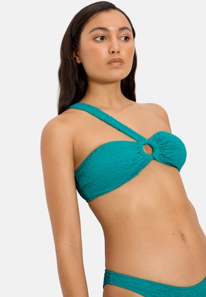 Mujer con un bikini texturizado color verde azulado, con una tiranta de un solo hombro y un recorte circular en el pecho, mirando hacia un lado.