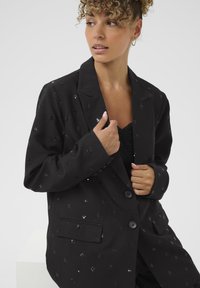 Schwarzer Blazer aus strukturiertem Stoff, mit kleinen Pailletten, Reverskragen und zwei Knöpfen. Enthält seitliche Taschendetails.