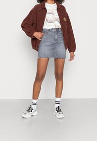 Žena v hnědé kordové bundě, grafickém bílém tričku, džínsové sukni s neopracovaným lemem, bílých ponožkách s černými pruhy a robustních teniskách.