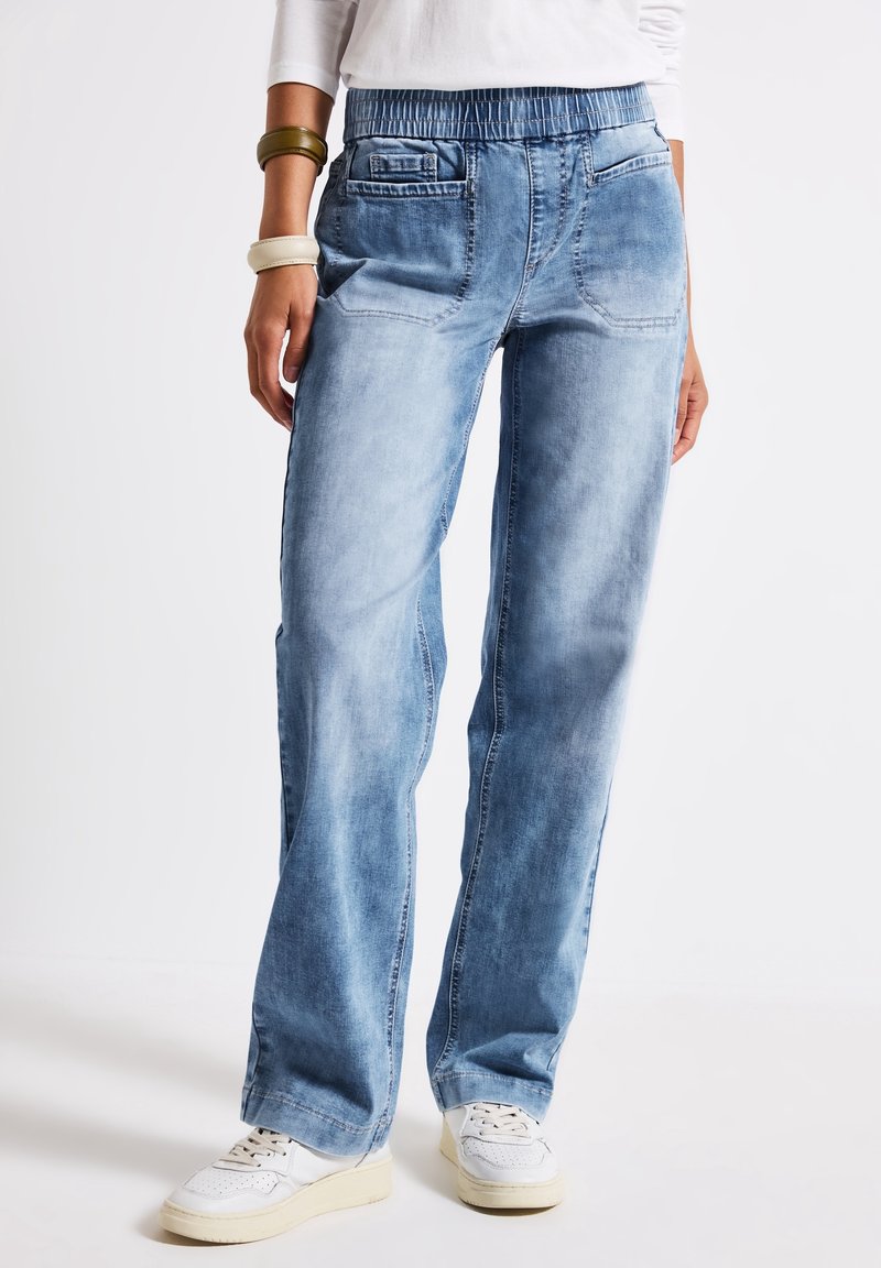 Femme portant un jean bleu clair à coupe ample avec une taille élastique et des baskets blanches, debout devant un fond clair uni.