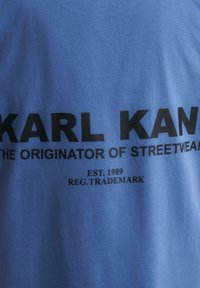 Mėlyna medvilninė marškinė su dideliu juodu tekstu: "KARL KANI," "GATVĖS MADOS KŪRĖJAS," "ĮSTEIGTA 1989," ir "REG. PREKĖS ŽENKLAS." Lygus paviršius.