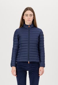 Marineblauwe pufferjack met verticale quiltontwerpen, hoge kraag en ritssluiting aan de voorkant, met contrasterende oranje ritssluiting en zijzakken.