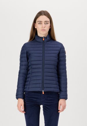 Marineblauwe pufferjack met verticale quiltontwerpen, hoge kraag en ritssluiting aan de voorkant, met contrasterende oranje ritssluiting en zijzakken.