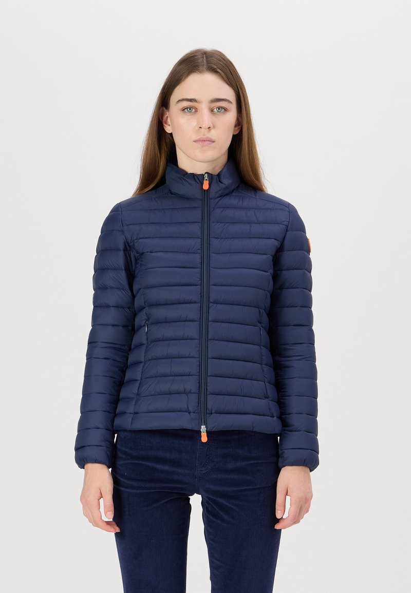 Marineblauwe pufferjack met verticale quiltontwerpen, hoge kraag en ritssluiting aan de voorkant, met contrasterende oranje ritssluiting en zijzakken.