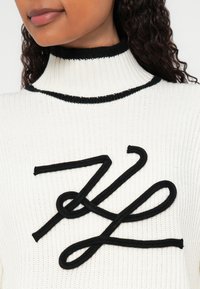 Beli pleteni pulover z črnim rebrastim ovratnikom in izrazitim črnim dekorativnim vrvnim vzorcem na sprednji strani.