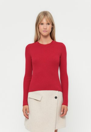 Roter, gerippter Langarmpullover mit taillierter Silhouette, kombiniert mit einem beigen Wickelrock, der eine Seitenasche und schwarze Knopfdetails aufweist.