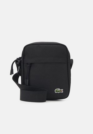 Bolso bandolera negro con un tejido suave, cierre de cremallera, correa ajustable y un pequeño logo de cocodrilo verde en la parte frontal.