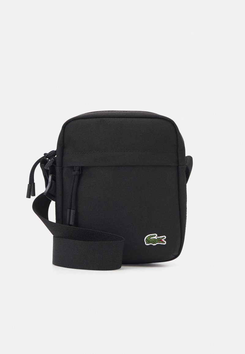 Bolso bandolera negro con un tejido suave, cierre de cremallera, correa ajustable y un pequeño logo de cocodrilo verde en la parte frontal.