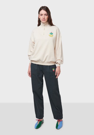Felpa di pile color crema con zip a metà, caratterizzata da ricami colorati. Pantaloni neri con tasca a toppa. Scarpe flat multicolori con lacci.