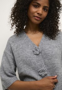 Pilka megzta kardiganas su V formos iškirpte, trumpomis rankovėmis ir dviem gėlių formos sagomis. Minkšta tekstūra su jaukia išvaizda.