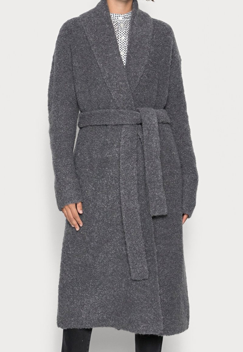 Manteau gris texturé mi-long de style ceinturé avec un large col et une ceinture assortie en tissu, porté sur un haut blanc à motifs et un pantalon noir.