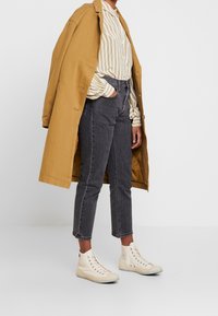 Cappotto lungo beige, camicia a righe gialla e bianca, jeans scuri e sneakers alte color crema. Vestibilità rilassata con texture a contrasto.